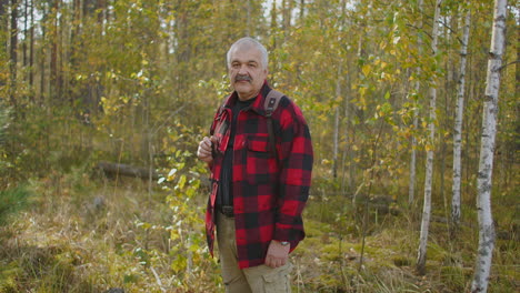 cheerful middle-aged man is standing in autumn forest with yellowed trees at daytime smiling to camera hiker and traveler