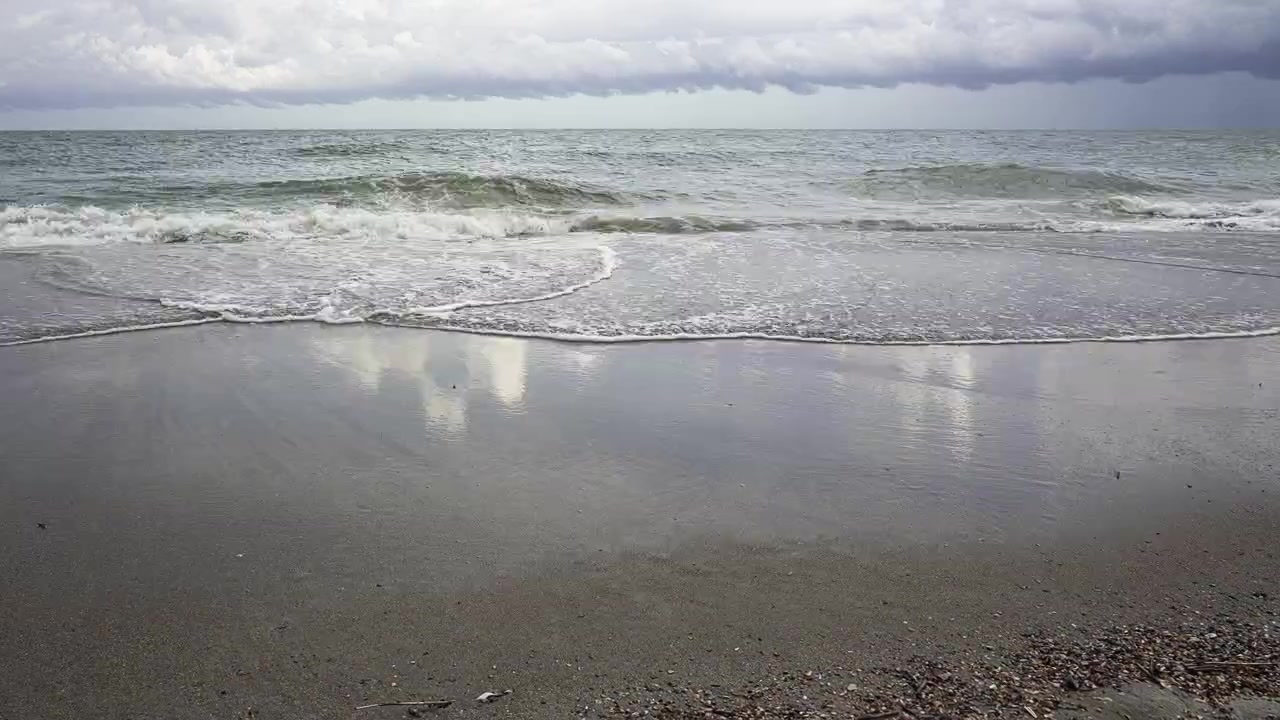 Fredfyldte havbølger ruller og skyller ind over en sandstrand under en skyet, overskyet himmel. Den rolige tidevand skaber en fredelig, naturlig scene i en time-lapse video
