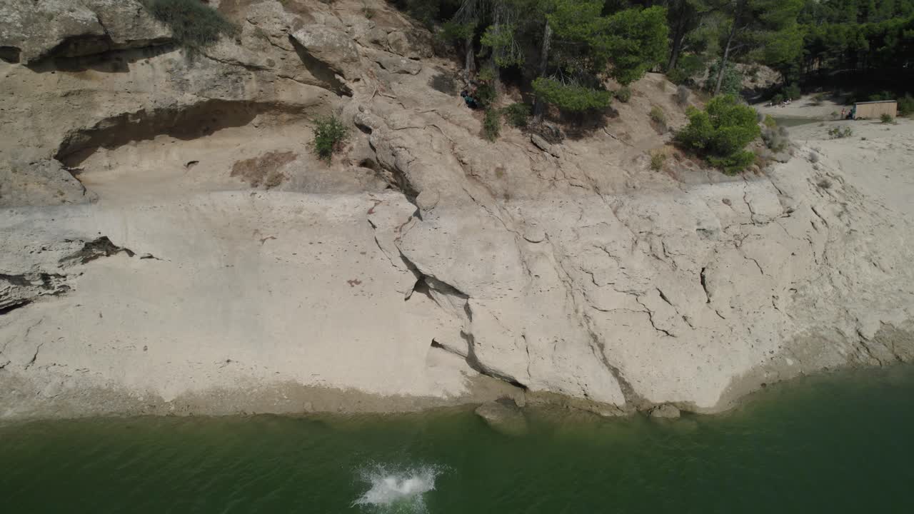 Panoramic drone shot of young man jumping into natural water body from edge of rock, with trees in background, enjoying holidays on solo trip to El Chorro of Málaga, Spain