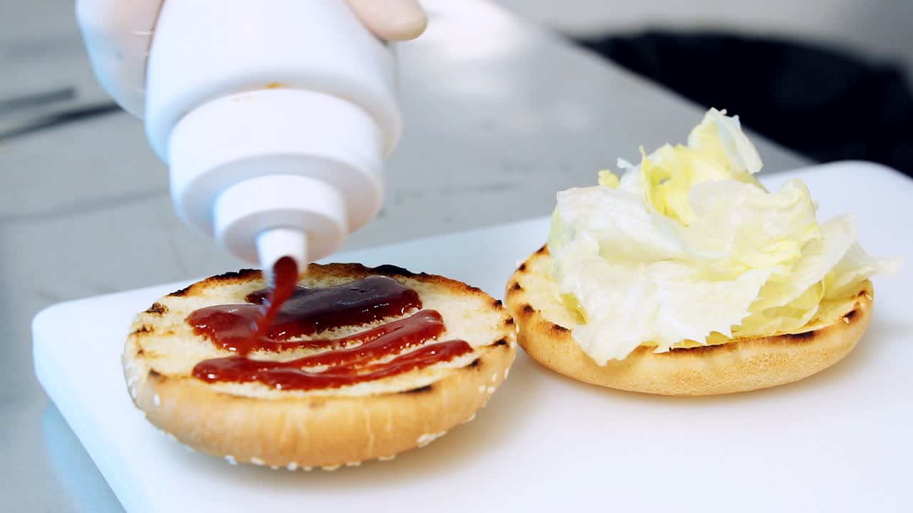 Chef pouring in fresh sweet ketchup on fried bun. Cooking delicious snack at table in kitchen. Chef spreads red sauce on a burger bun. Close-up