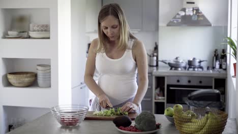 sonriente futura madre cortando ensalada en la mesa de la cocina