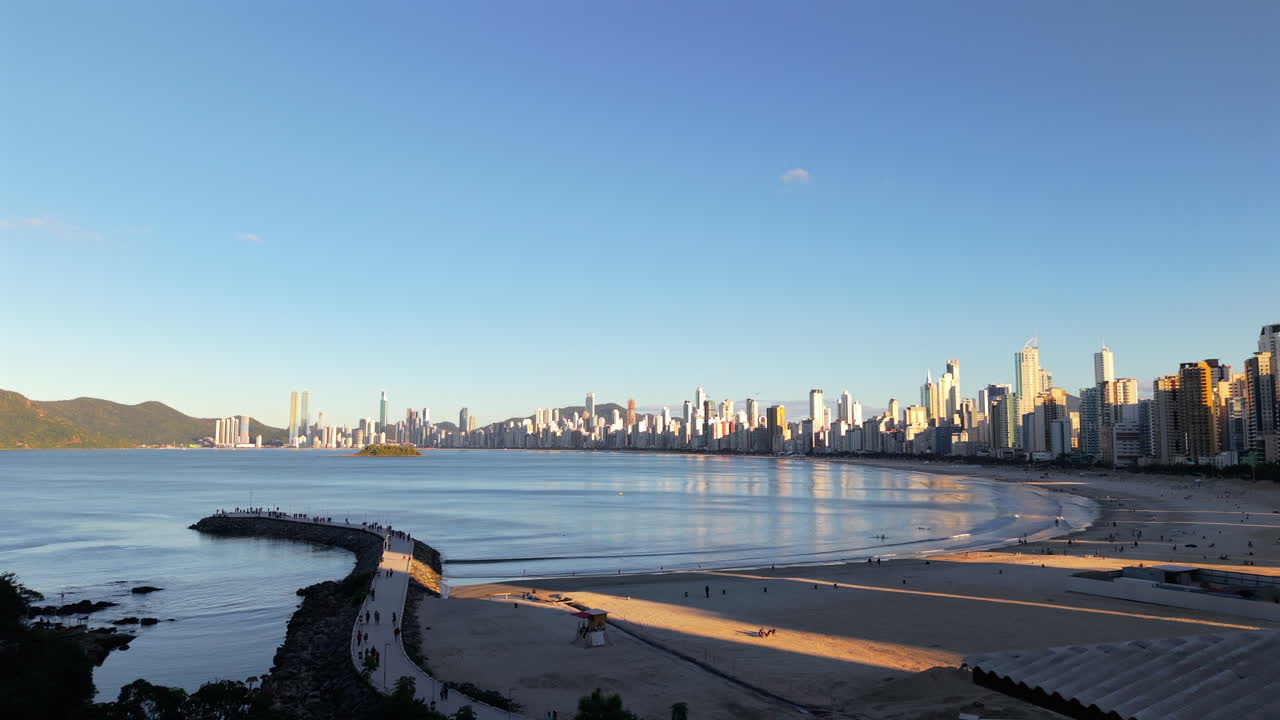 Drone sobrevoando a orla de Balneário Camboriú de trás para frente, com o cais de concreto, arranha-céus à direita e morros à esquerda. Vista geral. Tarde.
