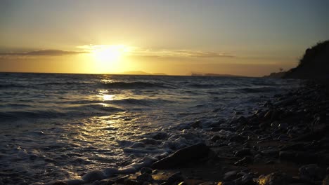 Slow-motion-shot-of-waves-crashing-on-the-coast-of-Corfu-with-the-sun-setting,-low-angle-view