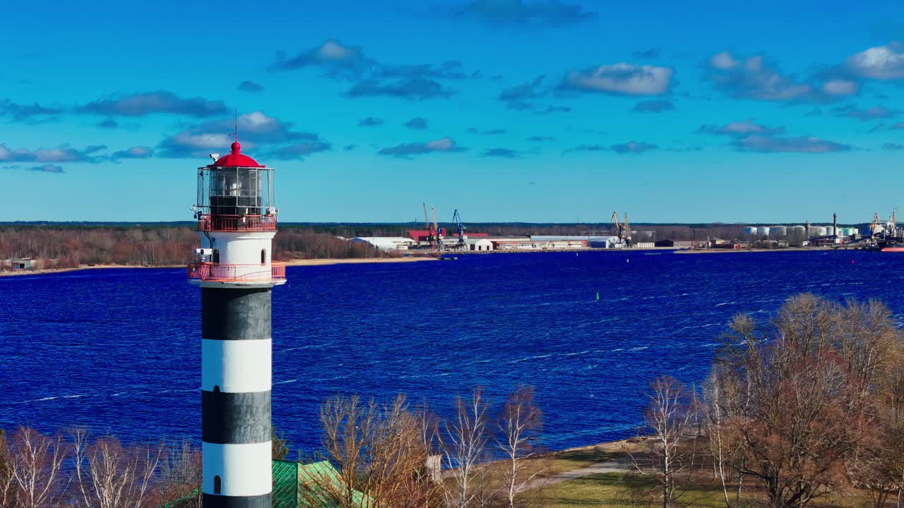 A tall black-and-white lighthouse capped with bright red stands beside deep blue water, framed by leafless trees and a distant shoreline under clear sky.