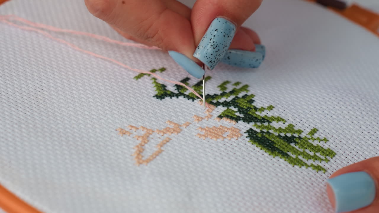 Close up of fashion designer embroidering delicate green leaf pattern on white fabric, needle moving with precision and care, blue polished nails highlighting craftsmanship