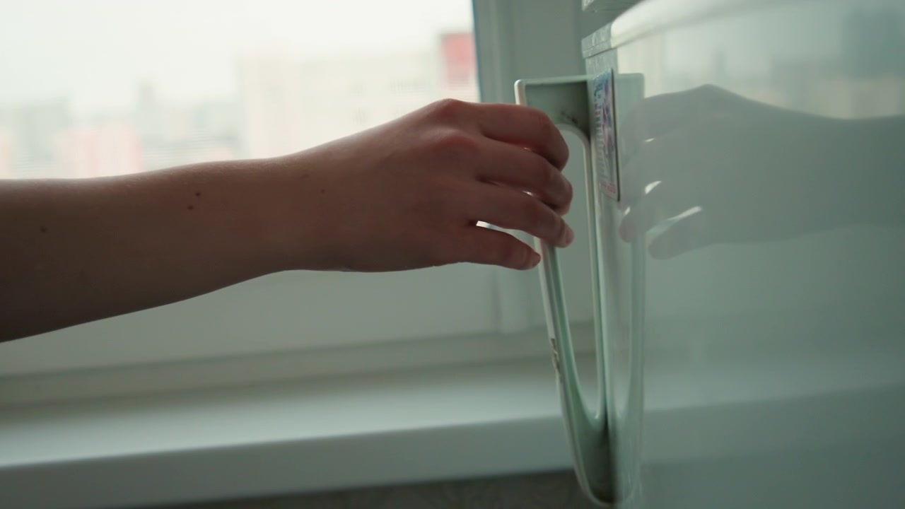 Hand reaching fridge handle before cooking, medium closeup of hand pulling stainless handle with magnet sticker visible, city window background, precise movement, evening prep for dinner, practical