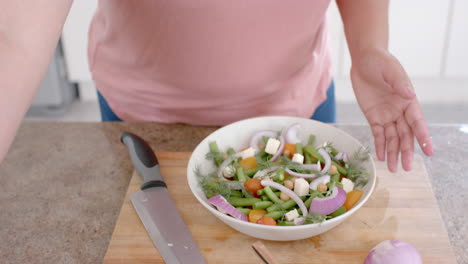 mujer biracial de tamaño plus feliz haciendo queso feta y ensalada de verduras en la cocina, en cámara lenta
