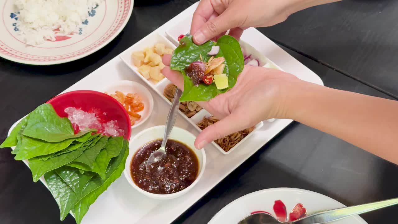 Hands assemble Miang Kham by adding fillings and sauce onto betel leaves, with vibrant ingredients arranged on a white platter under bright, even lighting