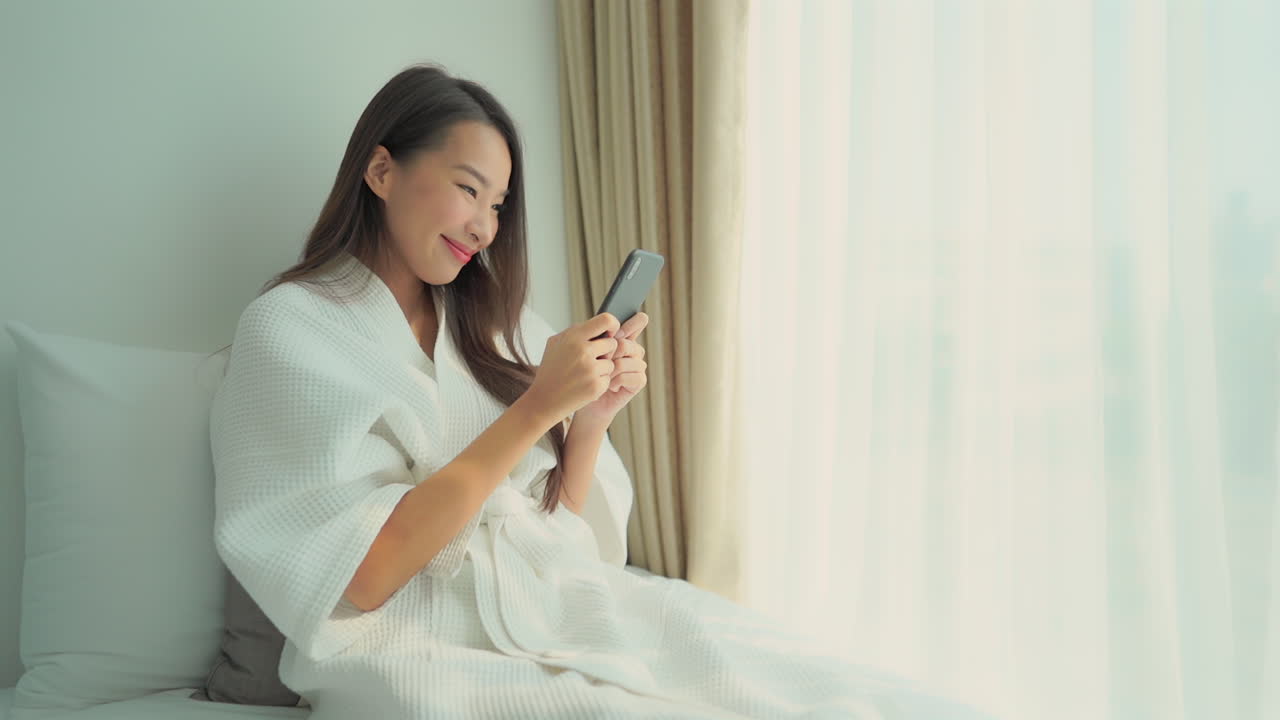 Nice Asian woman in bathrobe sits on bed, smiles and looks at mobile