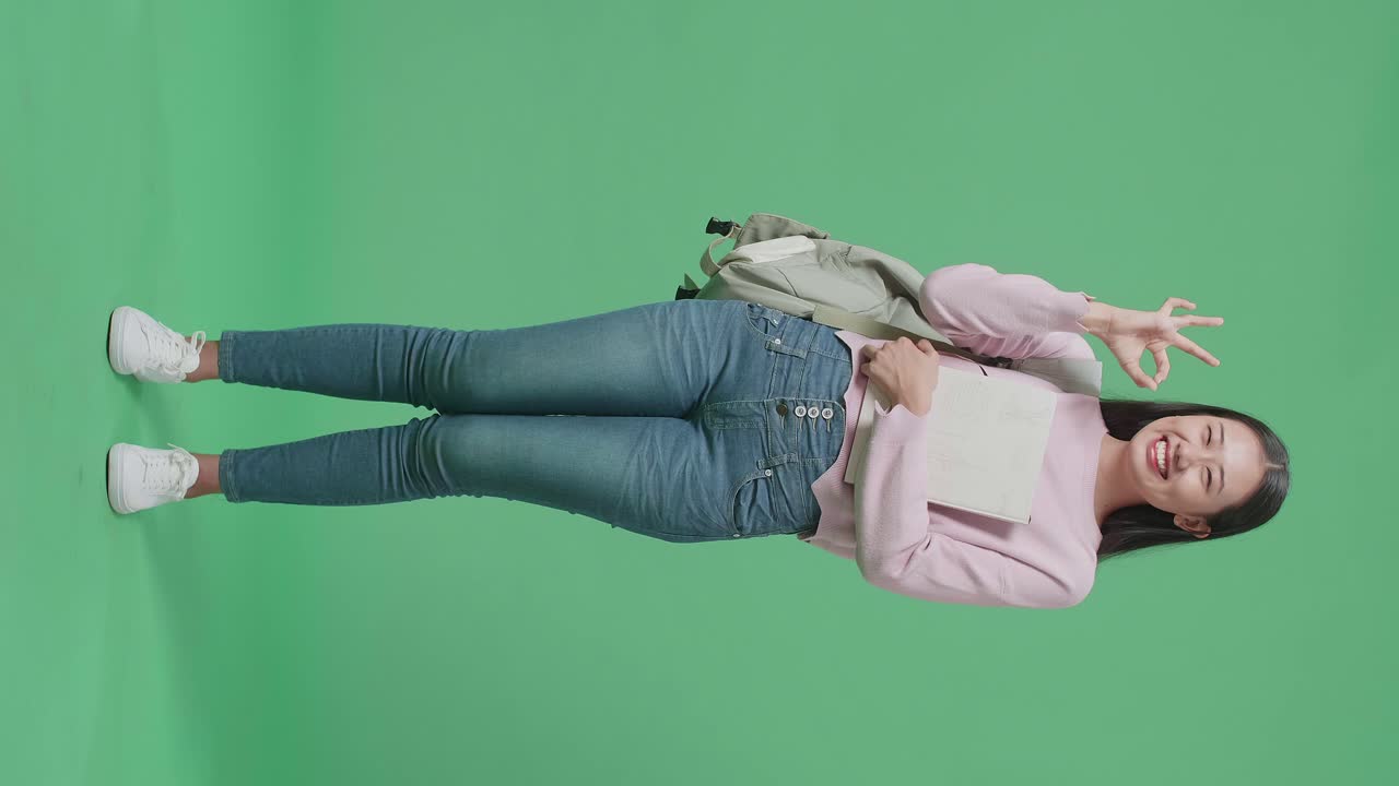 Full Body Of Asian Woman Student Holding Book And Showing Ok Gesture On Green Screen Studio
