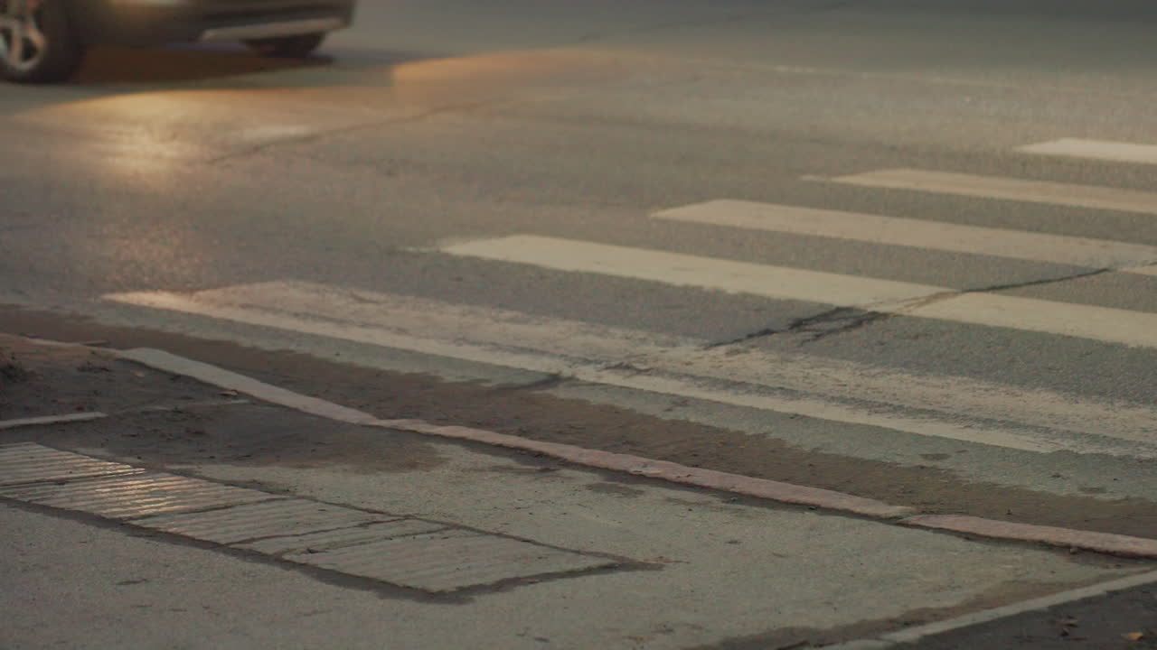 Close up view of car passing over pedestrian crosswalk with white stripes on rough asphalt at street junction during evening light, showing detailed urban road surface with lines