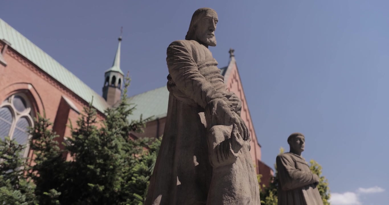 Dynamický záběr mramorové sochy svatého apoštola držícího dětskou ruku, s kostelem a stromy v pozadí za slunečného letního dne, natočeno ve 4K rozlišení a zpomaleném záběru.