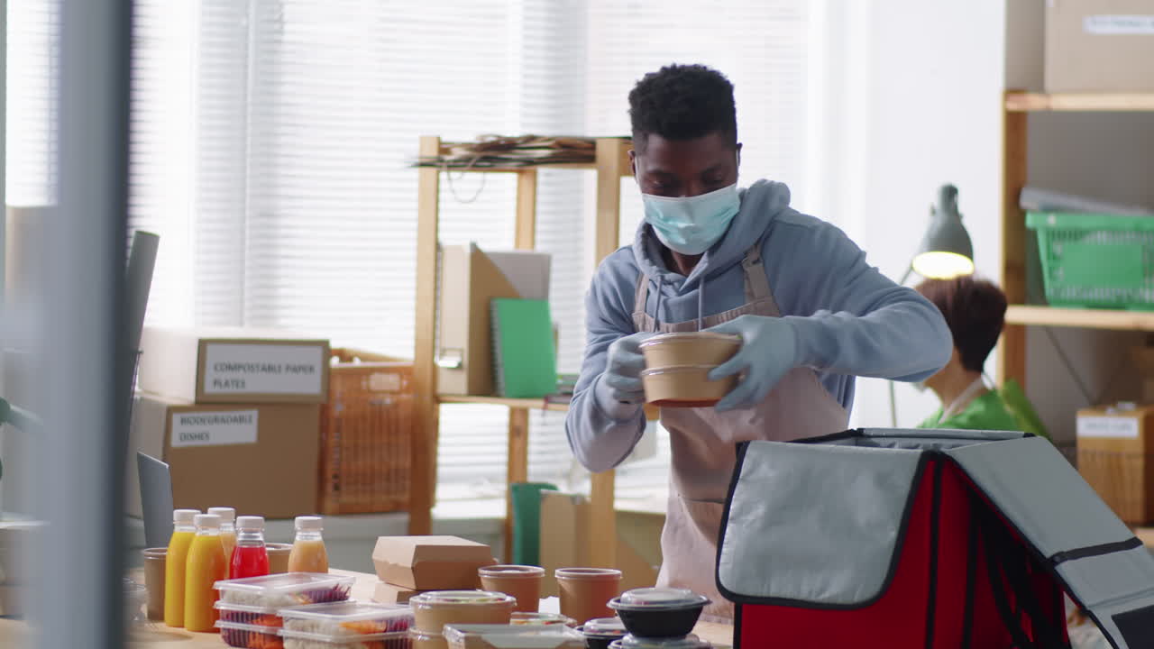 Black Man in Mask Packing Food Boxes and Drinks in Delivery Bag