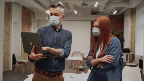 young woman and man with face masks and a laptop talk about work with in the office