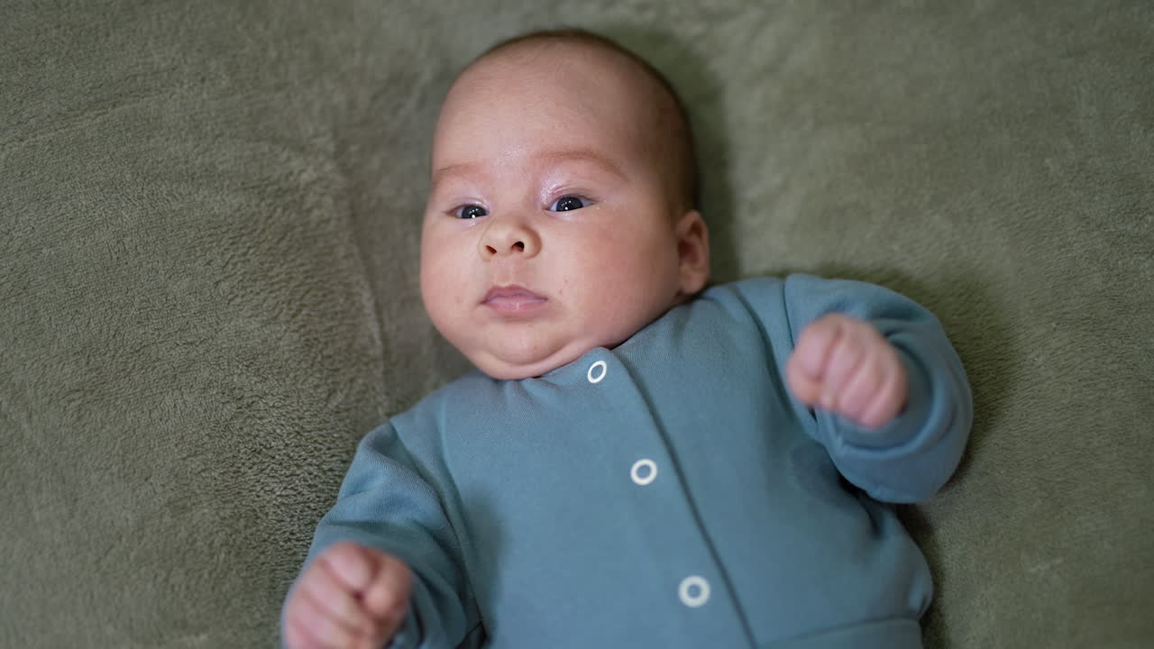 Cute baby boy with plump cheeks lying on the bed. Toddler in blue suit on the grey blanket. View from above.
