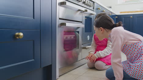 Two-Girls-Waiting-For-Cupcakes-To-Bake-Looking-Into-Oven-In-Kitchen-At-Home-Together