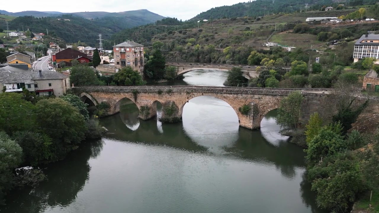 Ampia vista del Ponte da Cigarrosa che attraversa il fiume Sil tra Petín e A Rúa de Valdeorras, con i suoi storici archi in pietra riflessi nell'acqua calma e i villaggi che si ergono sullo sfondo in Galizia, Spagna.
