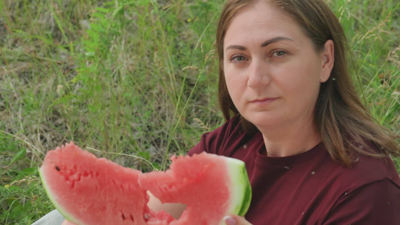 mujer caucásica sentada sosteniendo una rodaja de sandía, mirada tranquila y camisa color granate, fondo de campo de hierba, suave mordisco creando una abertura similar a una máscara, picnic informal en el campo, momento gastronómico espontáneo, iluminado por el sol