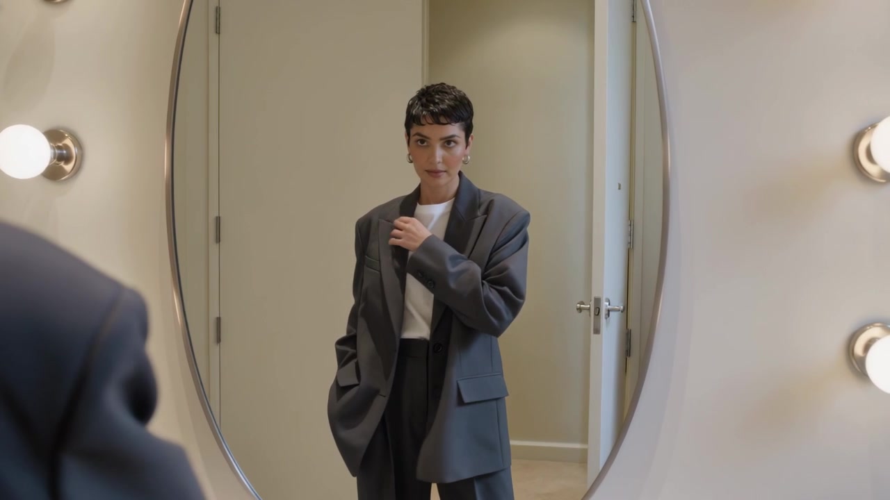 Donna che si muove in tailleur grigio, si sistema il risvolto del blazer nella zona spogliatoio, lo specchio attira lo sguardo.