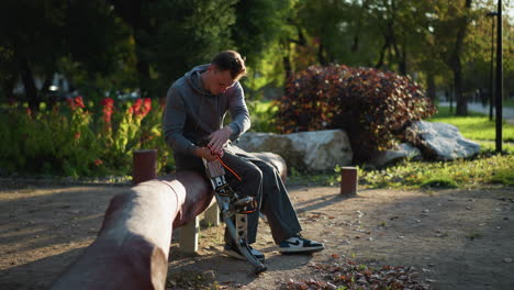 hombre de cabello marrón sentado en un tronco en el parque con una sudadera gris con capucha, pantalones gris oscuro, zapatillas nike air jordan 1, ajustando pilotes de primavera plateados y negros con expresión seria bajo árboles con luz solar