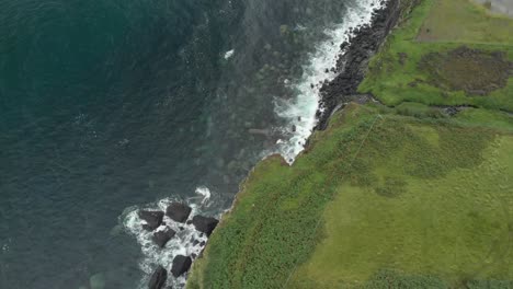 drone-shot-of-waterfall-ending-in-a-sea-cliff-in-isle-of-skye-scotland,-green-grass-and-clear-blue-ocean-water