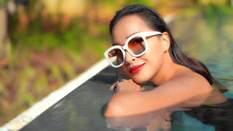 Cerca-De-Feliz-Mujer-Asiática-Sensual-En-Gafas-De-Sol-Disfrutando-Del-Sol-En-La-Piscina,-Cámara-Lenta