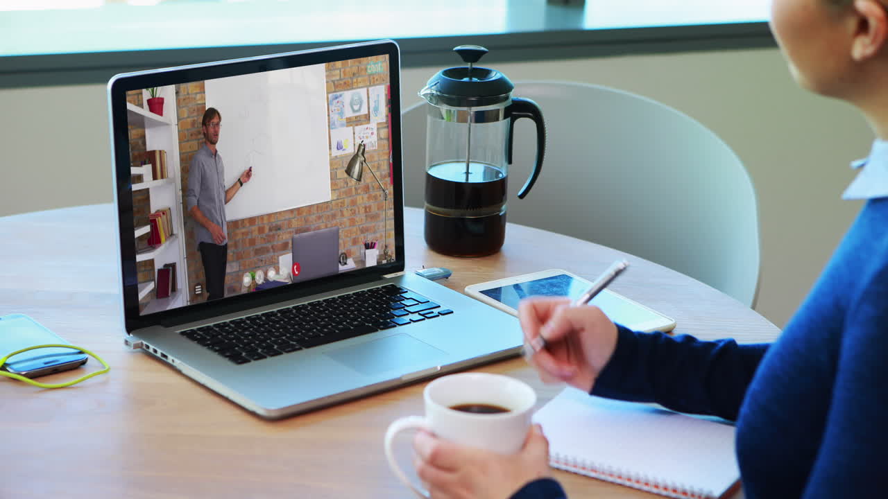 Caucasian female student using laptop on video call with male teacher, making notes