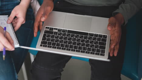 Black man with laptop on lap indoors closeup. African American employee uses modern computer sitting with colleague in office. Guy works online via device