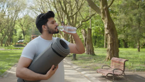 man drinking water in a park with yoga mat