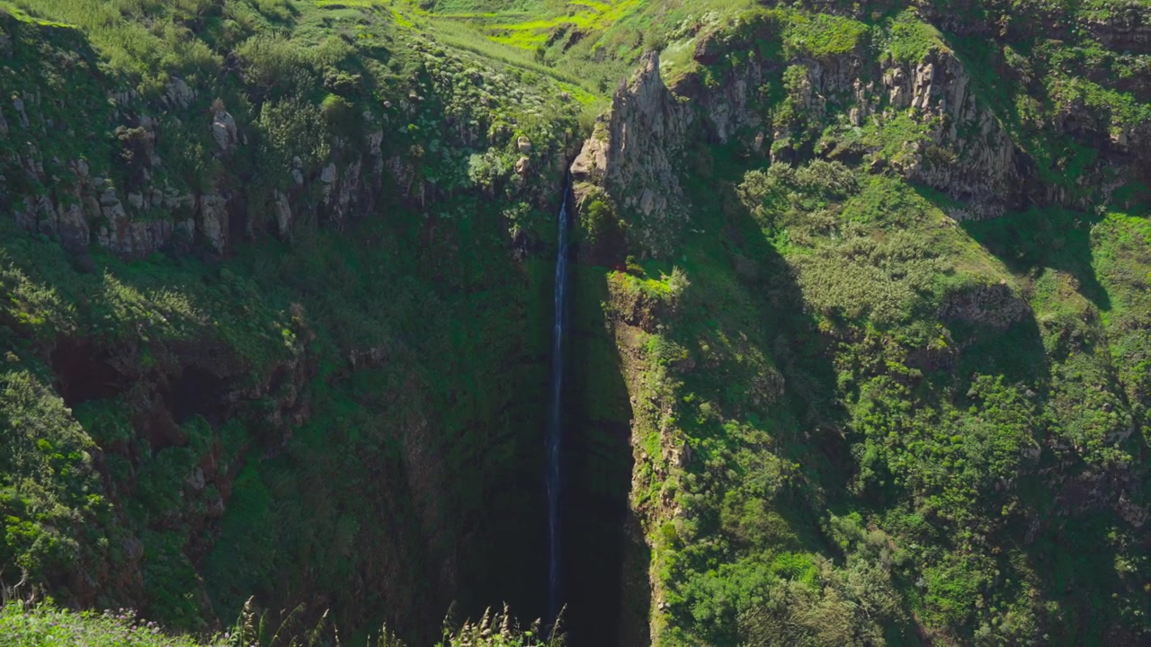 Una cascata alta e stretta scende a cascata da una ripida scogliera rocciosa, circondata da una lussureggiante vegetazione verde e terreno accidentato in pieno giorno - Ripresa a mano libera.