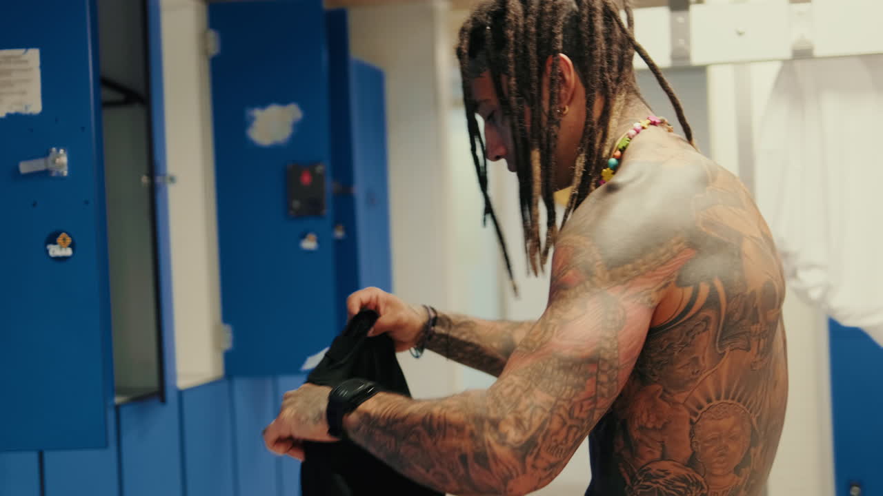 Tattooed Man with Dreadlocks Changing Clothes in a Locker Room