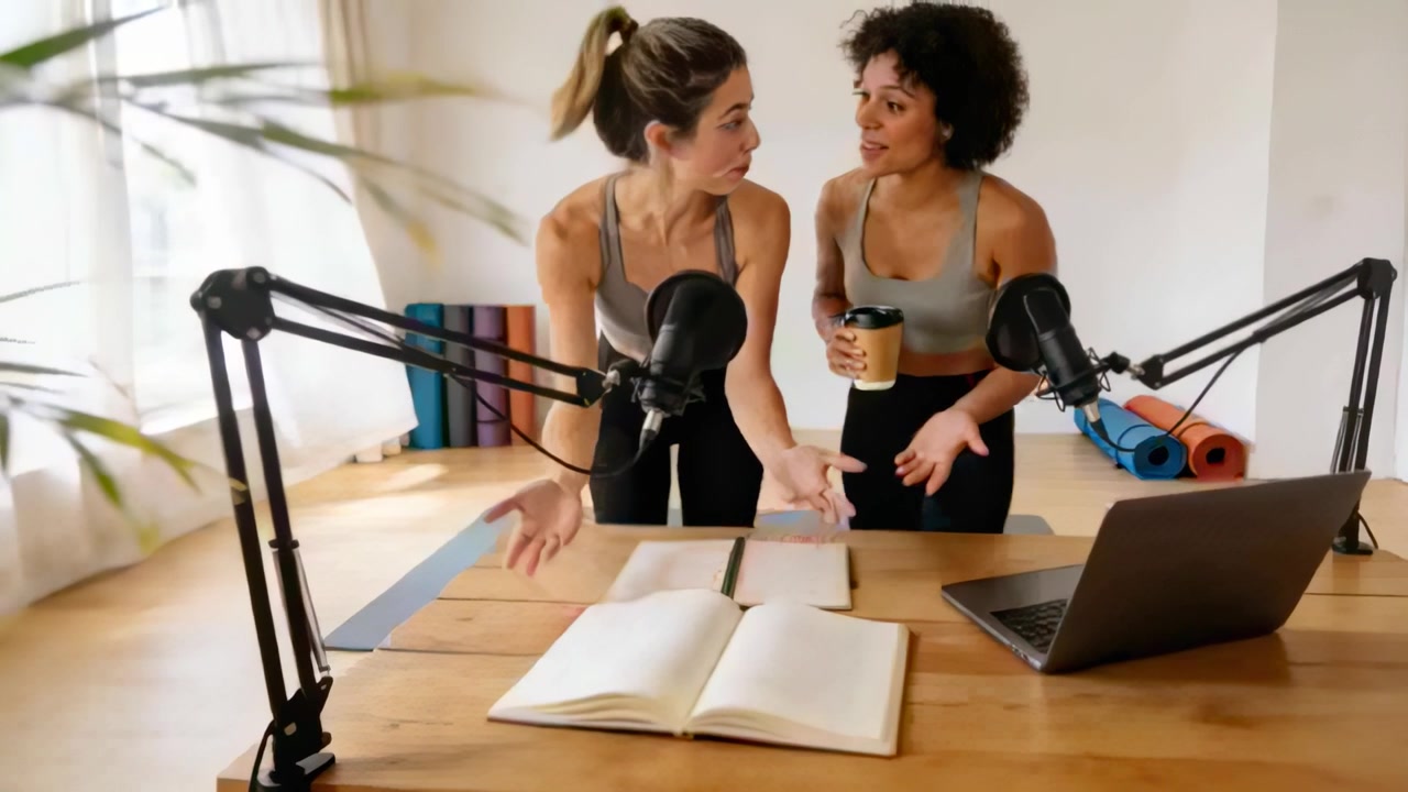 Coanfitriones con ropa deportiva inclinados hacia adelante, examinando un libro cerca de micrófonos y un portátil en el estudio mientras preparan notas.