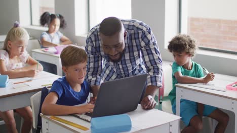 Video-of-happy-african-american-male-teacher-helping-caucasian-boy-with-laptop
