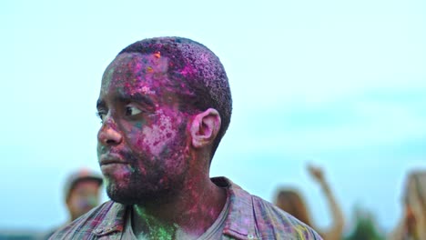 portrait of the young handsome man in colorful paints smiling to the camera and posing outdoors while having festive holi day with multiethnic friends