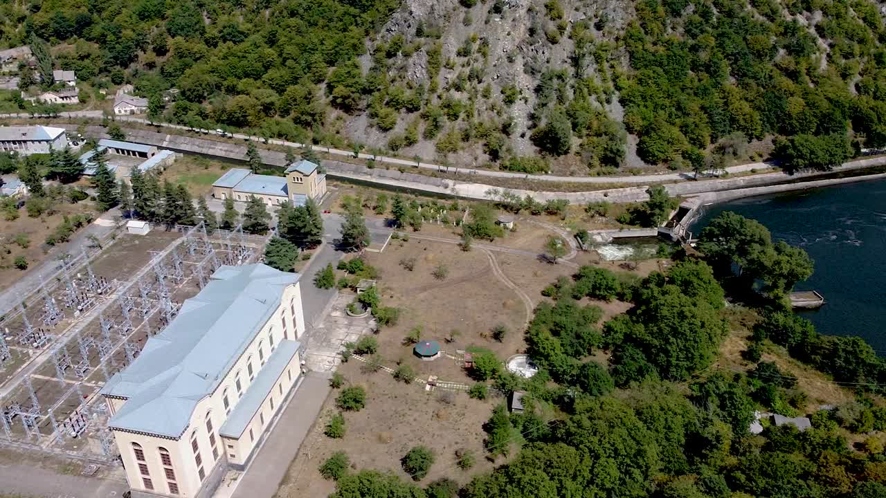 An aerial perspective showcases an industrial complex near a river, surrounded by greenery and mountain terrain. The area features buildings, power lines, and natural beauty