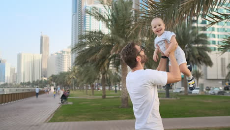 Father-playing-with-sons-in-the-summer-in-a-modern-city-holding-him-and-hugging-standing-in-a-white-t-shirt-and-shorts