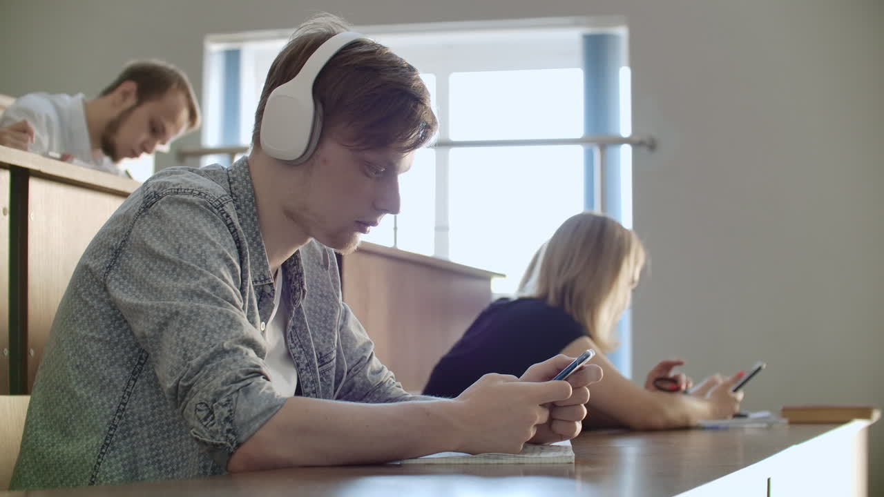 A student sitting in a university auditorium with a large group of people sits in headphones and listens to music and looks at the smartphone screen. It is in a state of calm. Class break
