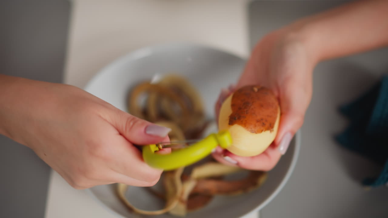 Close up view of fair skinned person peeling sweet potato using yellow peeler over bowl filled with potato skins, with blue kitchen rag slightly blurred on side of dining table