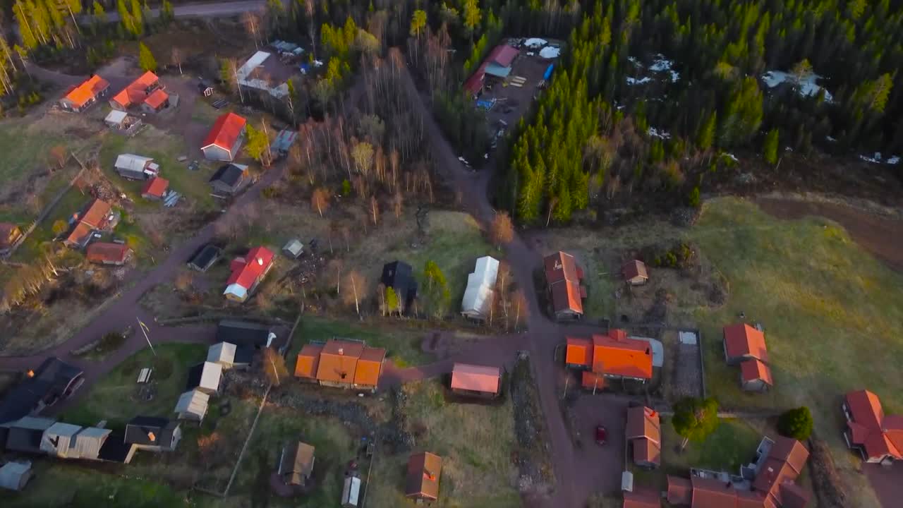 Aerial drone footage of a small housing area or a village with red rooftops in the middle of sunny Sweden pine forest nature, camera rises up and reveals the forest and a large lake in the background.