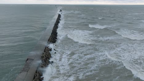 aerial establishing view of port of liepaja concrete pier , autumn storm, big waves splashing, overcast day, wide drone shot moving forward