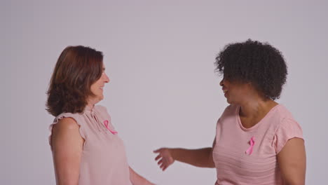 Studio-Portrait-Of-Two-Smiling-Women-Of-Different-Ages-Wearing-Pink-Clothing-And-Breast-Cancer-Awareness-Ribbons-Hugging-Against-White-Background