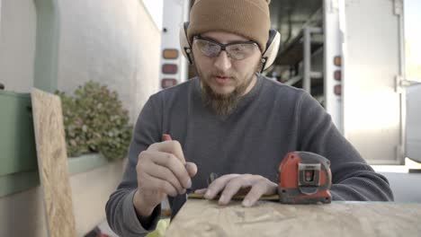 artesano con gafas de seguridad y orejeras marca la madera con lápiz y cinta métrica