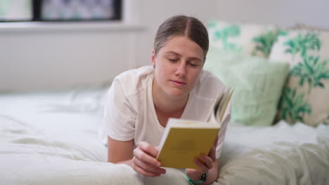 4k, joven mujer caucásica acostada en la cama leyendo un libro y pasando páginas