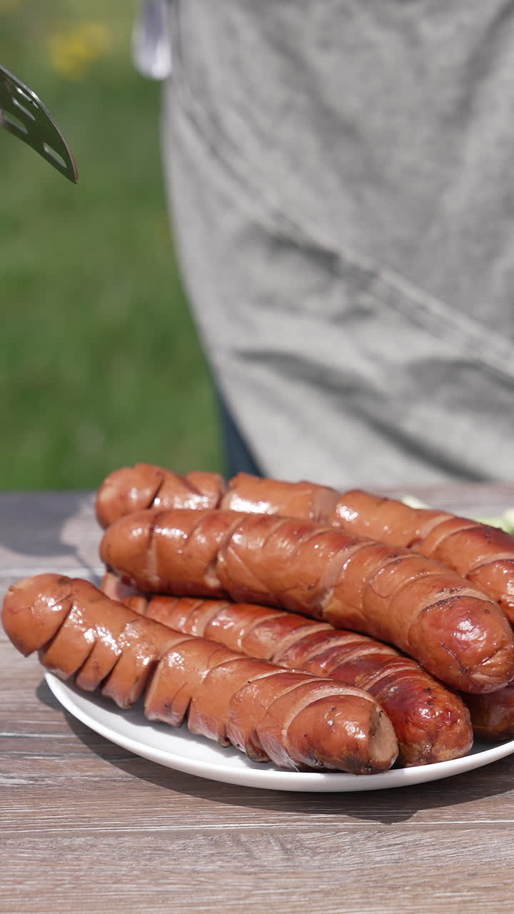 Ready grilling sausages with vegetables on the table outdoors. Chef puts roast sausage into a plate. Tasty meal for a picnic. Vertical video