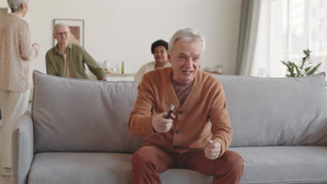 Medium long of senior Caucasian man sitting on couch in living room, watching TV emotionally, calling for his friend, sharing news
