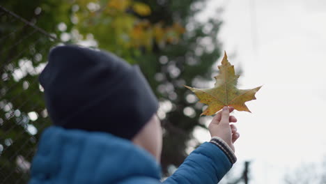 close-up of hand holding dried rusted leaf, rotating it thoughtfully, observing intricate details with blurred background of vibrant greenery and sunlight filtering through leaves