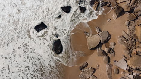 Bird-eye-aerial-view-of-ocean-waves-crashing-at-rocky-and-stony-beach-in-Nazare,-Portugal