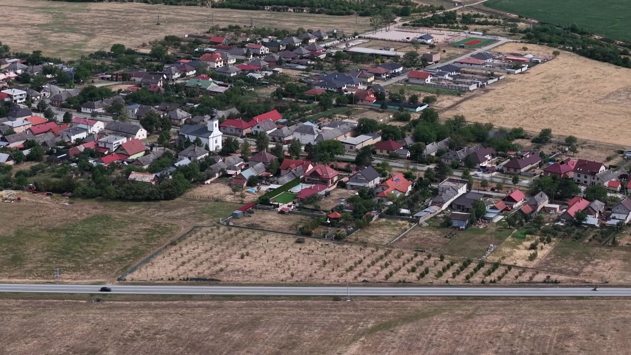 A high-angle aerial drone shot showcases a peaceful European village with red-roofed houses and a central church surrounded by fields.