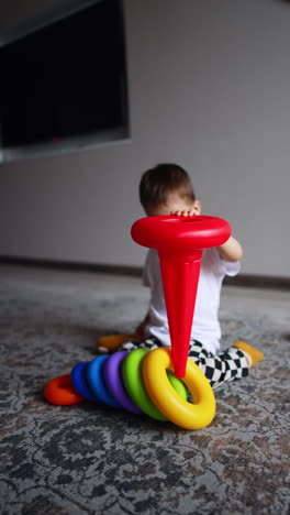 little toddler boy breaking the pyramid. kid holds the toy basis sitting on the floor in his room. happy kid playtime.