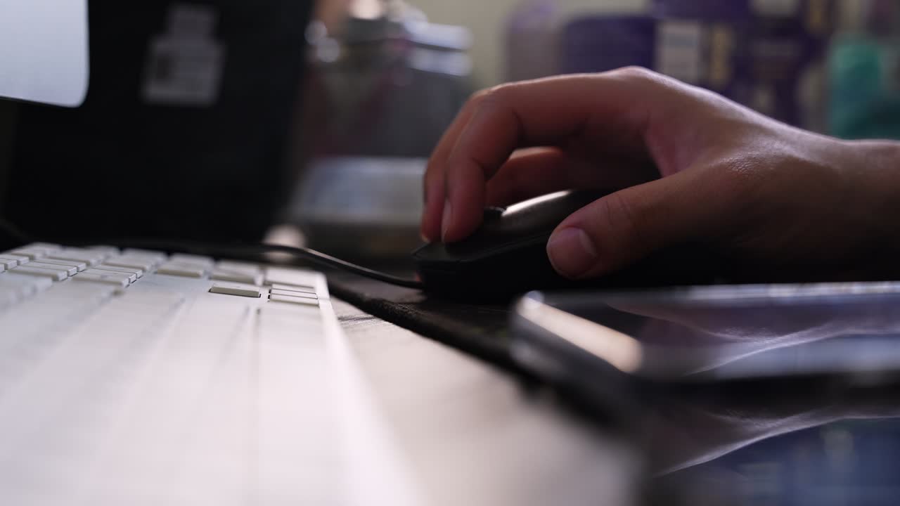 a hand operating a computer mouse beside a keyboard and smartphone on a desk, with soft indoor lighting, shallow focus, and an atmospheric workspace background